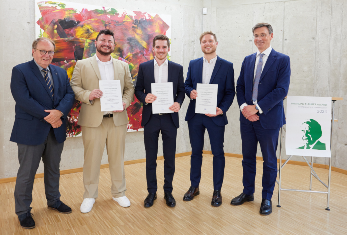 Fünf Männer posieren mit Urkunden vor einem abstrakten Gemälde und einem Schild.