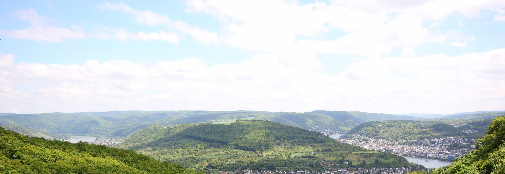 Landschaftsbild mit grünen Hügeln, einem Fluss und einer Stadt unter einem bewölkten Himmel.