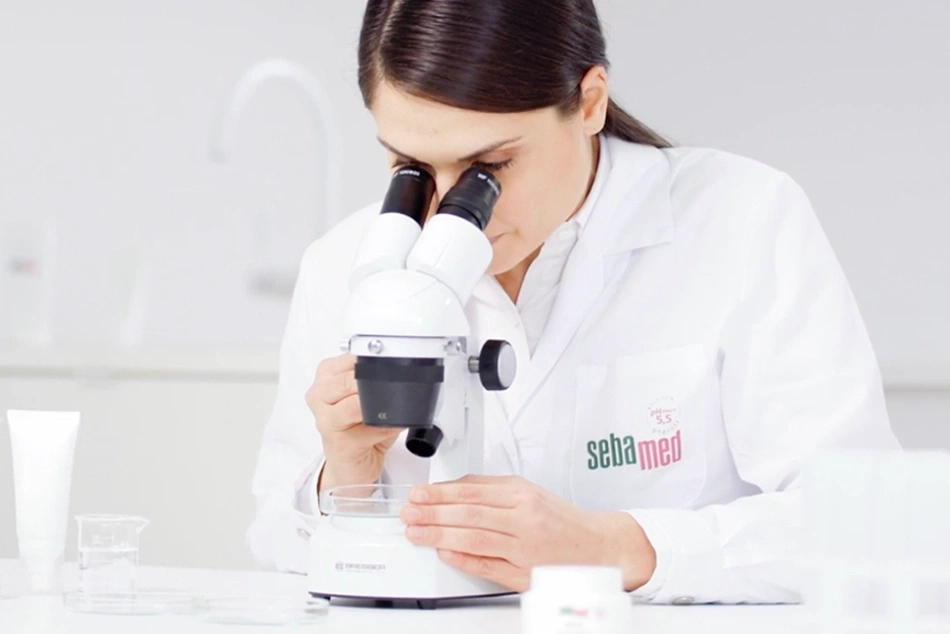 Wissenschaftlerin betrachtet Probe durch Mikroskop im Labor, SebaMed-Logo