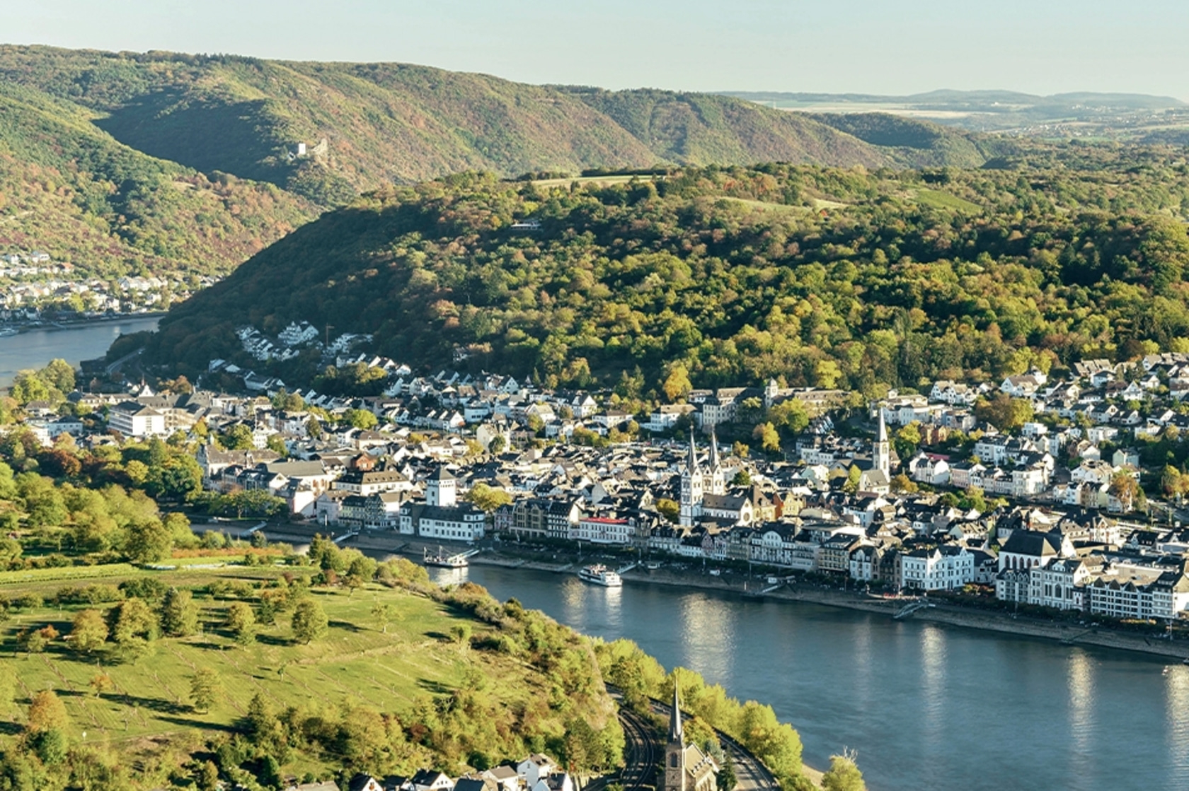 Luftaufnahme einer malerischen Stadt am Flussufer, umgeben von grünen Hügeln und Bäumen.