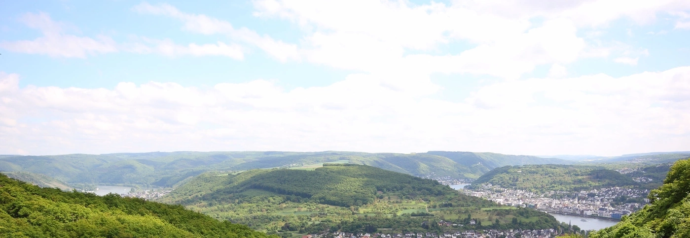 Landschaftsbild mit grünen Hügeln, einem Fluss und einer Stadt unter einem bewölkten Himmel.