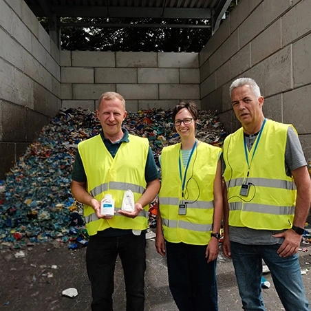 Drei Personen mit Warnwesten stehen vor einem Haufen recycelter Materialien. Links hält jemand zwei weiße Flaschen.