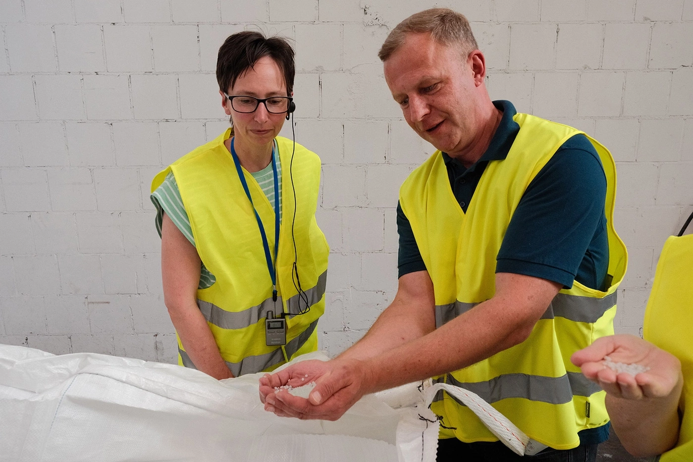 Zwei Personen in Warnwesten untersuchen weiße Granulatperlen in einem industriellen Umfeld.