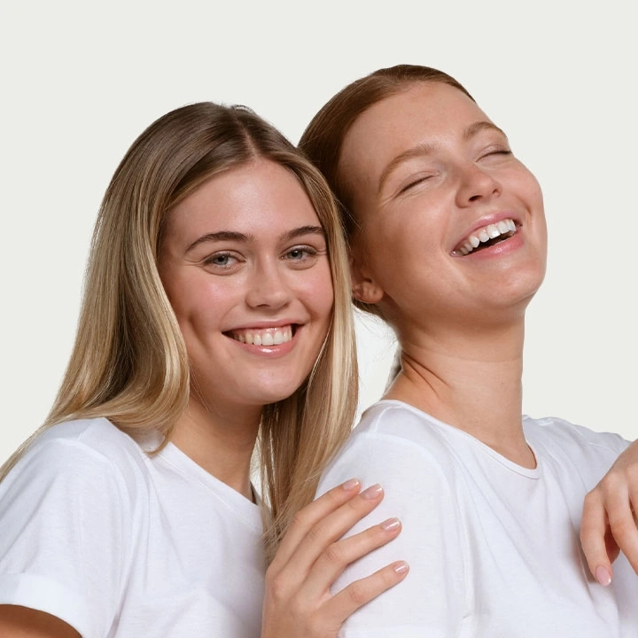Zwei lächelnde Frauen in weißen T-Shirts, die sich umarmen und lachen.